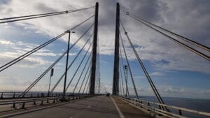 Eine malerische Aussicht auf eine Schrägseilbrücke, die sich über ein Gewässer erstreckt, aufgenommen aus einem fahrenden Fahrzeug. Der Himmel ist teilweise bewölkt, während die Sonne die Straße und die Seile beleuchtet und eine atemberaubende Kulisse schafft, perfekt für diejenigen, die von einer unvergesslichen Hochzeit träumen.