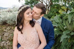 Ein Bräutigam im blauen Anzug küsst die Wange der Braut während ihrer Hochzeit. Die Braut, in einem rosa Kleid mit Blumendetails und einer Blumenkrone, lächelt sanft. Sie stehen in einem Garten mit üppigem grünem Laub, was eine ruhige und romantische Atmosphäre schafft, die perfekt für ihre Hochzeit ist.