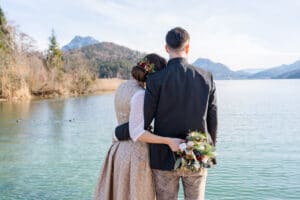 Ein Paar steht mit abgewandtem Blick an einem ruhigen See. Die Frau trägt ein Blumenhaarteil und einen Blumenstrauß in der Hand und ist in traditioneller Kleidung gekleidet. Es ist die perfekte Kulisse für ihre Hochzeit, mit Bergen und Bäumen, die die Szene unter einem klaren Himmel einrahmen.