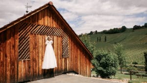 Ein weißes Brautkleid hängt an einer rustikalen Scheunenwand und verkörpert die Essenz einer Hochzeit. Im Hintergrund sind weitläufige grüne Weinberge unter einem bewölkten Himmel zu sehen, mit verstreuten Häusern zwischen den Hügeln, die die Romantik einfangen, die Hochzeitsplanung inspiriert.