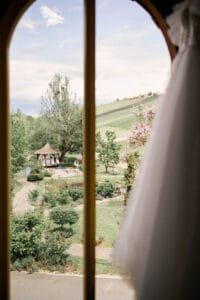 Der Blick durch ein Fenster zeigt einen malerischen Garten mit üppigem Grün, einem Pfad und einem bezaubernden Pavillon. Ein Hochzeitskleid hängt teilweise sichtbar auf der rechten Seite und deutet auf Hochzeitsplanung für eine malerische Hochzeit im Freien oder ein besonderes Ereignis hin.