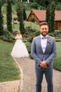 Eine Braut in einem weißen Kleid geht auf einen Bräutigam im blauen Anzug zu, der auf einem Gartenweg steht. Sie sind von Grün und rustikalen Gebäuden umgeben und bilden die perfekte romantische Kulisse für ihre Hochzeit – eine verträumte Umgebung im Freien, ideal für diejenigen, die sich in eine sorgfältige Hochzeitsplanung vertiefen.