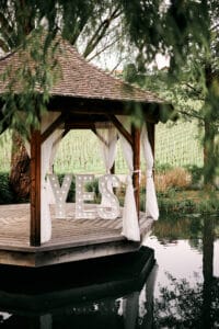 Ein hölzerner Pavillon mit weißen Vorhängen steht über einem Teich, umgeben von Grün, perfekt für eine Hochzeitszeremonie. Große beleuchtete Buchstaben mit der Aufschrift „JA“ sind im Pavillon angebracht und schaffen eine romantische Atmosphäre, ideal für diejenigen, die heiraten möchten.