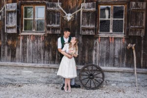Ein Paar in traditioneller Kleidung steht vor einer rustikalen Holzhütte, die die perfekte Kulisse für ihre Hochzeit darstellt. Mit Fensterläden und Geweihen darüber ist die Szene idyllisch für jede Hochzeitsplanung. Die Frau hält einen Blumenstrauß, während ein hölzernes Wagenrad an der Hütte lehnt.