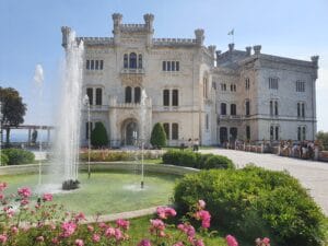 Ein großes, historisches Schloss mit kunstvoller Architektur steht unter einem klaren blauen Himmel, perfekt für diejenigen, die ihre Traumhochzeit planen. Im Vordergrund ist ein großer Brunnen von blühenden rosa Blumen und üppigen grünen Sträuchern umgeben. In der Nähe des Eingangs sind Menschen zu sehen, die die Aussicht genießen.