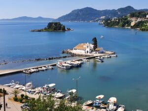 Eine malerische Aussicht auf eine Küstenregion, perfekt für eine romantische Hochzeit. Mit einem kleinen weißen Gebäude auf einem Pier, umgeben von Booten und klarem blauen Wasser. Dahinter sind eine üppig grüne Insel und in der Ferne Hügel unter dem klaren blauen Himmel zu sehen.