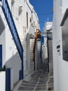 Eine schmale Gasse in einer griechischen Inselstadt, perfekt für Hochzeitsfotos, zeichnet sich durch weiße Gebäude mit leuchtend blauen und braunen Treppengeländern aus. Der klare, blaue Himmel verstärkt den hellen Kontrast der weißen Wände und der dunkel gefärbten Geländer.