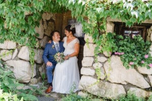 Braut und Bräutigam sitzen zusammen unter einem Baldachin aus grünen Blättern, perfekt eingebettet in eine rustikale Steinmauer. In ihrem freudigen Moment der Hochzeit hält die Braut einen Blumenstrauß in der Hand und blickt den Bräutigam in seinem blauen Anzug an, während sie die Schönheit genießt, die die Natur zu ihrer Hochzeit bringt.
