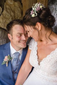 Braut und Bräutigam teilen einen freudigen Moment an ihrem Hochzeitstag. Die Braut, in einem weißen Spitzenkleid mit Blumen im Haar, lehnt sich zum Bräutigam hinüber, der einen blauen Anzug und eine Blumenboutonniere trägt. Sie lächeln und stehen eng beieinander und genießen die Freude des Heiratens.
