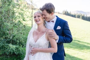 Eine Braut in einem weißen Spitzenkleid und Schleier lächelt, während ihr Partner im blauen Anzug sie von hinten umarmt. Sie stehen in einer grasbewachsenen Umgebung mit sonnendurchfluteten Bäumen und Hügeln und fangen die Magie ihres Hochzeitstages ein.