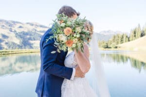 Braut und Bräutigam stehen umarmt vor einem ruhigen See und beginnen ihre Hochzeitsreise. Der Brautstrauß bedeckt elegant ihre Gesichter. Majestätische Berge und üppige Bäume zieren den Hintergrund, alles gespiegelt im ruhigen Wasser unter einem klaren blauen Himmel.