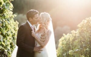Braut und Bräutigam küssen sich in einem sonnenbeschienenen Weinberg und verkörpern damit die Essenz einer Hochzeit. Die Braut trägt einen langen, fließenden Schleier und ein funkelndes Kleid, während der Bräutigam einen dunklen Anzug trägt. Üppige grüne Weinreben umgeben sie, während das Sonnenlicht an ihrem besonderen Tag der Hochzeit einen sanften, romantischen Schein verbreitet.