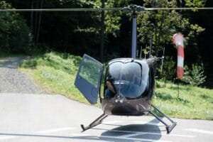 Ein Hubschrauber mit offener Tür steht auf einem Landeplatz in einem Waldstück und ist bereit für eine einzigartige Hochzeitseinfahrt. Im Hintergrund flattert ein Windsack, der dezent die Windrichtung für diesen unvergesslichen Moment des Heiratens anzeigt.