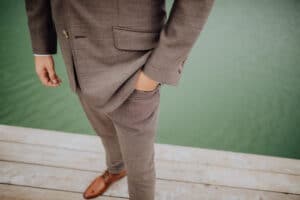 Eine Person in einem braunen Anzug, die vielleicht über Hochzeitspläne nachdenkt, steht mit einer Hand in der Tasche auf einem Holzsteg und blickt auf grünes Wasser. Ihr Kopf ist im Bild nicht sichtbar und sie trägt braune Schuhe.