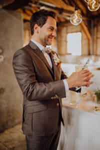 Ein Mann im grauen Anzug hält ein mit Rosmarin garniertes Getränk in der Hand und lächelt, er genießt die Hochzeitsfeier sichtlich. Er steht in einem warm beleuchteten Raum mit Holzbalken und hängenden Glühbirnen, sein Hochzeitsboutonniere ist perfekt an seiner Jacke befestigt.