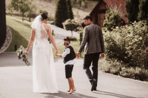 Eine Braut in weißem Kleid und Schleier und ein Bräutigam im Anzug gehen Hand in Hand mit einem kleinen Jungen, der Sonnenbrille und Weste trägt und die Freude an der Hochzeitsplanung verkörpert. Sie sind an einem sonnigen Tag im Freien mit einer malerischen Kulisse aus Grün und einem rustikalen Gebäude.