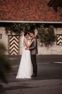 Eine Braut und ein Bräutigam, tief verliebt, teilen einen intimen Moment im Freien inmitten des Charmes ihrer Hochzeit. Sie strahlt in einem weißen Kleid mit Schleier, während er in einem dunklen Anzug gut aussieht. Hinter ihnen trägt ein rustikales Gebäude mit gemusterten Türen zur bezaubernden Hochzeitskulisse bei.