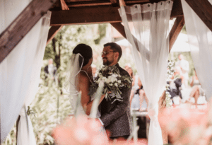 Braut und Bräutigam stehen in einer bezaubernden Hochzeitsszene unter einem rustikalen Holzpavillon, umgeben von hauchdünnen weißen Vorhängen. Die Braut hält einen Strauß weißer Blumen in der Hand, während sie sich liebevoll anschauen und in ihren perfekten Moment inmitten verschwommenen Grüns und Blüten versunken sind.