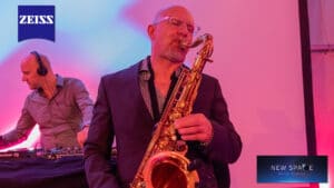 Auf der Bühne spielt ein Mann in einem dunklen Anzug leidenschaftlich Saxophon und verleiht der Atmosphäre gefühlvolle Töne, perfekt für jede Hochzeitsfeier. Im Hintergrund bedient ein anderer Mann mit Kopfhörern das DJ-Pult. Die Szene ist in bunte Lichter getaucht, während Logos von Zeiss und New Space Mission Research zu sehen sind.