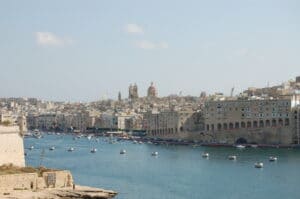 Eine Hafenszene mit ruhigem blauem Wasser und zahlreichen kleinen Booten in Valletta, Malta, bietet eine perfekte Kulisse für eine Traumhochzeit. Das Stadtbild zeigt historische Gebäude und Kirchen unter einem klaren blauen Himmel, links sind Befestigungsanlagen zu sehen – ideal für Hochzeitsplanung in einer zauberhaften Umgebung.