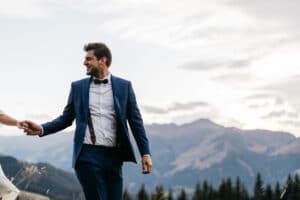 Ein Mann in blauem Anzug und Fliege lächelt, während er außerhalb des Bildes mit einer Person Händchen hält, die möglicherweise ihre Hochzeit feiert. Er steht in einer Berglandschaft unter einem bewölkten Himmel, mit baumbedeckten Hügeln im Hintergrund.