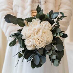 Eine Person in einem hellen Outfit hält einen Strauß weißer Rosen und Grünpflanzen, darunter Eukalyptusblätter, ideal für eine elegante Hochzeit. Die Umgebung strahlt formelle Eleganz aus, perfekt für alle, die sich mit Hochzeitsplanung beschäftigen.