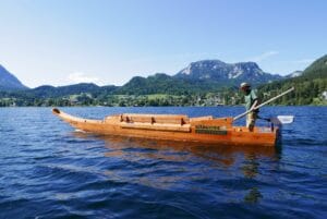 Eine Person steht und rudert ein traditionelles Holzboot auf einem ruhigen See, perfekt für Hochzeitspläne, umgeben von Bergen und üppigem Grün unter einem klaren blauen Himmel. Das Boot trägt die Beschilderung „Attersee Schifffahrt“ an der Seite.