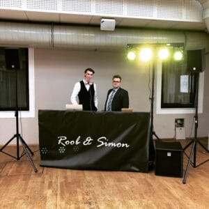 Zwei DJs stehen hinter einem Pult mit der Aufschrift „Rool & Simon“ in einem Raum mit Holzboden und sind bereit, Ihre Hochzeit unvergesslich zu machen. Umgeben von Lautsprechern und einer leuchtenden Lichtanlage mit grünem Licht schaffen sie die perfekte Atmosphäre für ein unvergessliches Fest der Liebe.