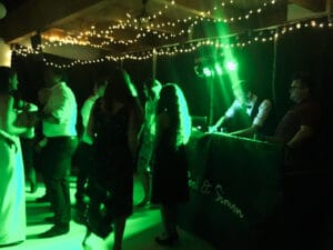Eine Gruppe von Menschen tanzt in einem schwach beleuchteten Raum mit grüner Beleuchtung und feiert eine unvergessliche Hochzeit. Von der Decke hängen Lichterketten, die einen magischen Schein verbreiten, während der DJ am „Cool & Simon“-Pult alle auf Trab hält und diesen Moment zu einem integralen Bestandteil ihrer Hochzeitsplanung macht.