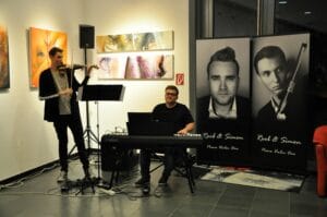 Zwei Musiker spielen im Innenbereich einer Hochzeit. Einer spielt Geige, der andere Keyboard. Hinter ihnen hängen Banner mit Porträts zweier Männer, die zur Atmosphäre dieses besonderen Anlasses beitragen. An der Wand hängen Kunstwerke, und in der Nähe steht ein Lautsprecher, um das intime Erlebnis zu verstärken.