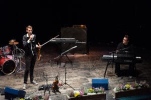 Zwei Musiker auf der Bühne: einer steht mit Mikrofon und Geige, der andere sitzt an einem Keyboard. Im Hintergrund sind ein Schlagzeug und ein zweites Keyboard zu sehen. Die mit Blumen geschmückte Bühne strahlt eine warme Atmosphäre aus, perfekt für unvergessliche Hochzeitsmomente.