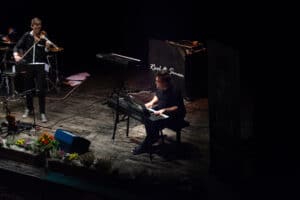 Auf einer schwach beleuchteten Bühne spielt ein Musiker Keyboard, links davon ein Geiger. Hinter ihnen steht ein „Rool & Simon“-Banner, das der Atmosphäre Eleganz verleiht. Die Bühne, die vorne mit bunten Blumen geschmückt ist, schafft die perfekte Stimmung für eine unvergessliche Hochzeitsfeier.