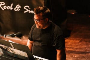 Eine Person mit Brille und schwarzem Hemd spielt auf der Bühne Keyboard und sorgt so für die perfekte Stimmung für eine Hochzeit. Ein Mikrofon steht in der Nähe und im Hintergrund ist ein teilweise sichtbares Schild mit den Worten „Rool & Sim“ zu sehen. Das Licht ist warm und auf den Künstler gerichtet.