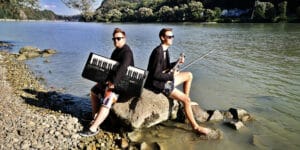 Zwei Personen in Anzügen sitzen auf felsigem Gelände an einem Fluss. Einer hält ein Keyboard, der andere eine Geige. Beide tragen Sonnenbrillen und Shorts und verbinden Hochzeitsplanung mit einem Hauch von Laune. Im Hintergrund ist eine malerische Aussicht mit Bäumen und Hügeln zu sehen, perfekt für eine unkonventionelle Hochzeitskulisse.