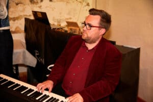 Ein Mann in einer roten Jacke spielt mit konzentriertem Gesichtsausdruck Klavier und perfektioniert vielleicht ein Stück für eine bevorstehende Hochzeit. Die rustikale Wand und der mit einem Laptop geschmückte Tisch lassen auf die komplizierten Details der laufenden Hochzeitsplanung schließen.