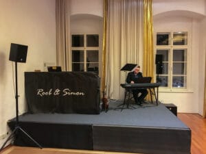 Ein Musiker spielt auf einer kleinen Bühne mit Lautsprechern und einem Ständer mit der Aufschrift „Rool & Simon“ auf einem Keyboard. Der warm beleuchtete Raum, der sich ideal für eine elegante Hochzeit eignet, verfügt über goldene und cremefarbene Vorhänge und einen Holzboden und schafft eine gemütliche Atmosphäre, die perfekt für jede Hochzeitsplanung ist.