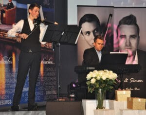 Zwei Musiker stehen bei einer Hochzeitsfeier auf der Bühne. Einer spielt Geige neben einem Notenständer, während der andere an einem Yamaha-Keyboard sitzt. Hinter ihnen hängt ein großes Banner mit ihren Fotos, was zur festlichen Atmosphäre beiträgt. Weiße Blumen in einer Vase schmücken die Vorderseite des Keyboards.