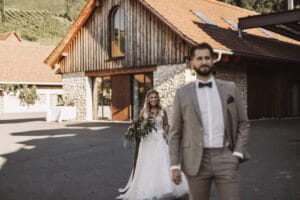 Eine Braut in einem weißen Kleid hält einen Blumenstrauß in der Hand und geht auf einen Bräutigam im grauen Anzug zu. Sie stehen vor einem rustikalen Gebäude aus Holz und Stein, das Sonnenlicht wirft lange Schatten auf den Boden – ein perfekter Moment, eingefangen in ihrer sorgfältig gestalteten Hochzeit.