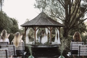 Ein Paar heiratet in einem rustikalen Holzpavillon mit weißen Vorhängen, umgeben von üppigem Grün. Gäste, Teil der sorgfältigen Hochzeitsplanung, sitzen auf Holzbänken im Vordergrund und beobachten die herzliche Zeremonie.