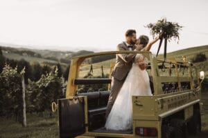 Braut und Bräutigam küssen sich auf dem Rücksitz eines gelben Oldtimers und verkörpern damit die Essenz einer perfekten Hochzeit. Die Braut hält ihren Brautstrauß hoch, während sie von einer malerischen Landschaft mit sanften Hügeln und Grün unter einem verblassenden Abendhimmel umgeben sind.