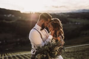 Ein Paar, das sich bei Sonnenuntergang umarmt, in einer malerischen Umgebung mit sanften Hügeln im Hintergrund, fängt die Essenz ihres Hochzeitstages ein. Die Frau hält einen Blumenstrauß, während sanftes Sonnenlicht diesen wunderschönen Moment in einen warmen Schein taucht.