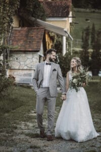 Braut und Bräutigam gehen Händchen haltend auf einem bezaubernden Weg im Freien spazieren. In seinem grauen Anzug mit Fliege und ihrem zarten weißen Spitzenkleid mit einem Blumenstrauß lächeln sie einander an. Das rustikale Gebäude und das üppige Grün bilden die perfekte Kulisse für ihr Hochzeitsabenteuer.