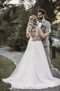 Ein Paar umarmt sich draußen auf einem Gartenweg und verkörpert seinen Hochzeitstraum. Die Frau trägt ein weißes Spitzenhochzeitskleid mit langer Schleppe, während der Mann einen hellgrauen Anzug mit Fliege trägt. Sie sind von üppigem Grün und einer ruhigen, natürlichen Kulisse umgeben – eine perfekte Szene für diejenigen, die heiraten möchten.