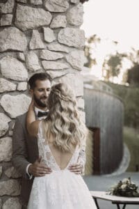 Ein Paar umarmt sich liebevoll neben einer Steinmauer auf seiner Hochzeit. Die Braut mit langen, welligen Haaren und einem Spitzenkleid blickt ihrem Partner entgegen. Der Bräutigam im Anzug lächelt sie an. Im Hintergrund sind ein rustikales Gebäude und Grünflächen zu sehen, die von sanftem Sonnenlicht beleuchtet werden.