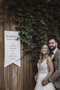 Ein glückliches Paar im Hochzeitskleid steht vor einer mit grünem Efeu geschmückten Holzwand. Neben ihnen definiert ein dekoratives Schild die Ehe humorvoll als „eine endlose Pyjamaparty mit deinem liebsten Spinner“. Es ist eine perfekte Momentaufnahme ihrer Reise von der Hochzeitsplanung bis zum Eheglück.