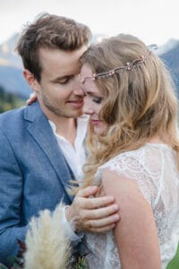 Ein Paar umarmt sich zärtlich im Freien und genießt einen Moment inmitten der Hochzeitsplanung. Der Mann im blauen Anzug hält die Frau, die ein weißes Spitzenkleid und ein mit Edelsteinen verziertes Haaraccessoire trägt, sanft im Arm, während sie eng beieinander stehen und eine ruhige Berglandschaft ihr Heiratsversprechen umrahmt.