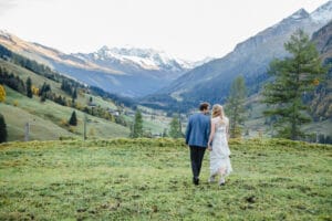 Ein Paar geht Hand in Hand über eine Wiese in Richtung der schneebedeckten Berge in der Ferne und verkörpert damit die Freude am Heiraten. Sie sind formell gekleidet, die Frau trägt ein weißes Kleid und der Mann einen Anzug. Die ruhige Szene ist von üppigem Grün und vereinzelten Bäumen geprägt, perfekt für die Hochzeitsplanung.
