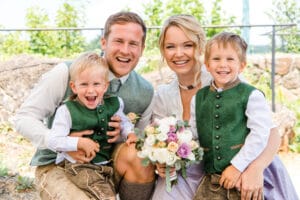 Eine lächelnde Familie posiert im Freien in traditioneller Kleidung und hält einen liebgewonnenen Moment inmitten der Aufregung der Hochzeit fest. Die Eltern knien und halten ihre beiden kleinen Jungen in grünen Westen und braunen Shorts. Die Mutter hält einen Strauß Rosen und anderer Blumen in der Hand, üppiges Grün bildet die perfekte Kulisse für ihre Feier.