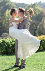 Ein Bräutigam im grauen Anzug hebt glücklich seine Braut im weißen Spitzenbrautkleid hoch. Sie hält einen Blumenstrauß in der Hand und feiert ihre Hochzeit im Freien auf einem üppigen grünen Rasen mit Bäumen im Hintergrund.