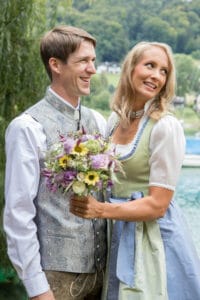Ein Paar in traditioneller bayerischer Kleidung steht an einem See und hält einen Moment seiner Hochzeit fest. Der lächelnde Mann hält einen Strauß Wildblumen, während die Frau lächelnd zur Seite schaut. Bäume und ein Gewässer bilden eine bezaubernde Kulisse für ihren besonderen Tag.